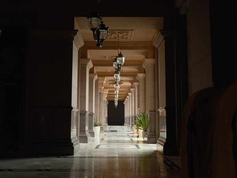 A dimly lit corridor with columns and hanging lamps, featuring a potted plant at the end.