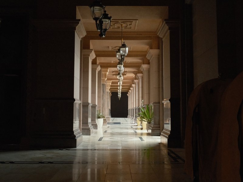 A dimly lit corridor with columns and hanging lamps, leading to a dark end with a potted plant.