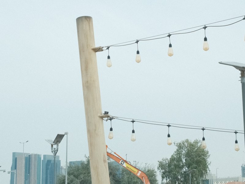 A wooden post with string lights against a cloudy sky, with buildings and construction equipment in the background.