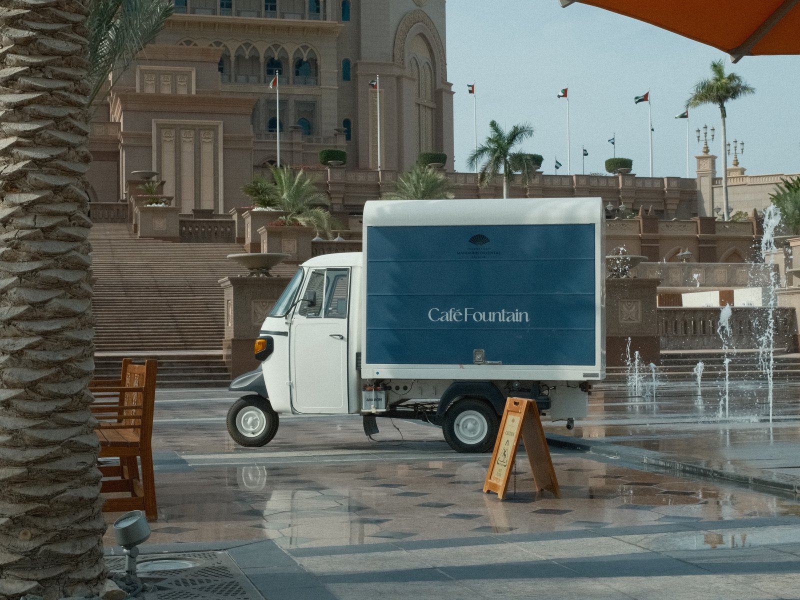 A small delivery truck parked near a fountain in a plaza with palm trees and a large building in the background.