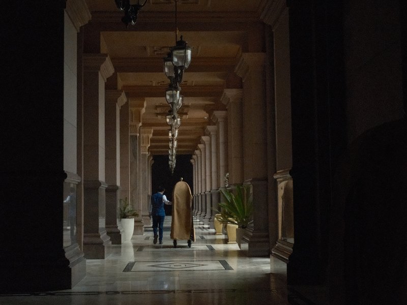 Two figures walking through a dimly lit corridor with decorative columns and hanging lanterns.