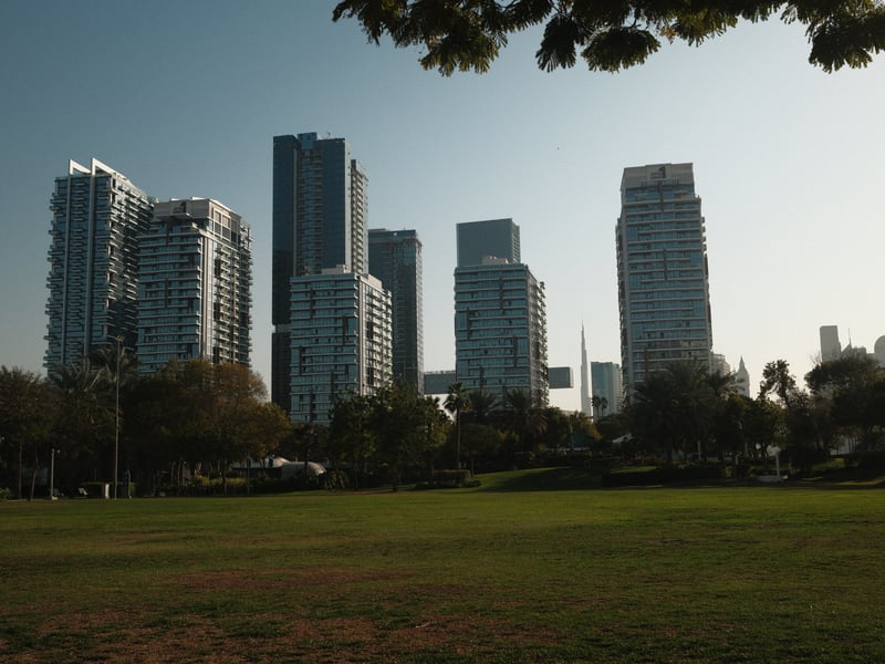 A skyline of modern skyscrapers viewed from a green park under a clear sky.