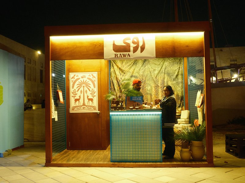 A small wooden stall at night with a sign, plants, and two people interacting inside.