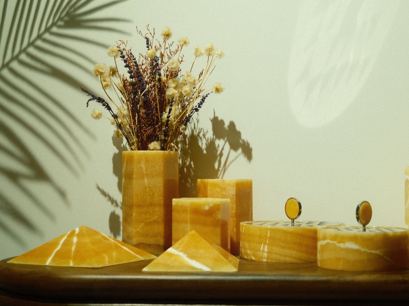 A still-life arrangement featuring yellow stone geometric shapes and a vase with dried flowers against a wall.