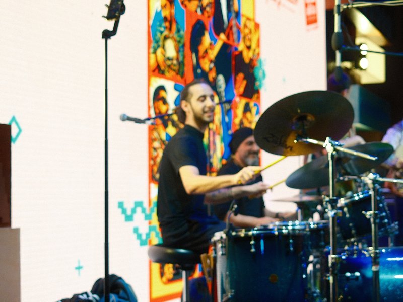 A drummer performing on stage with colorful artwork in the background, smiling while playing.