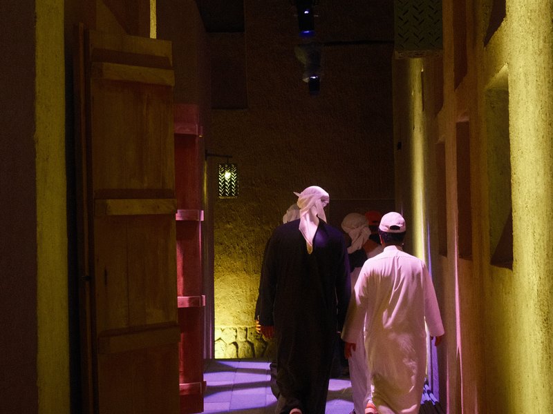 Two men walking through a narrow, illuminated corridor with traditional architecture.