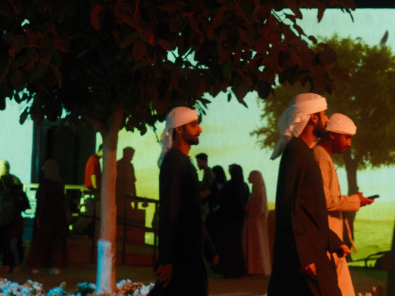 Group of people walking in traditional attire against a colorful backdrop with trees.