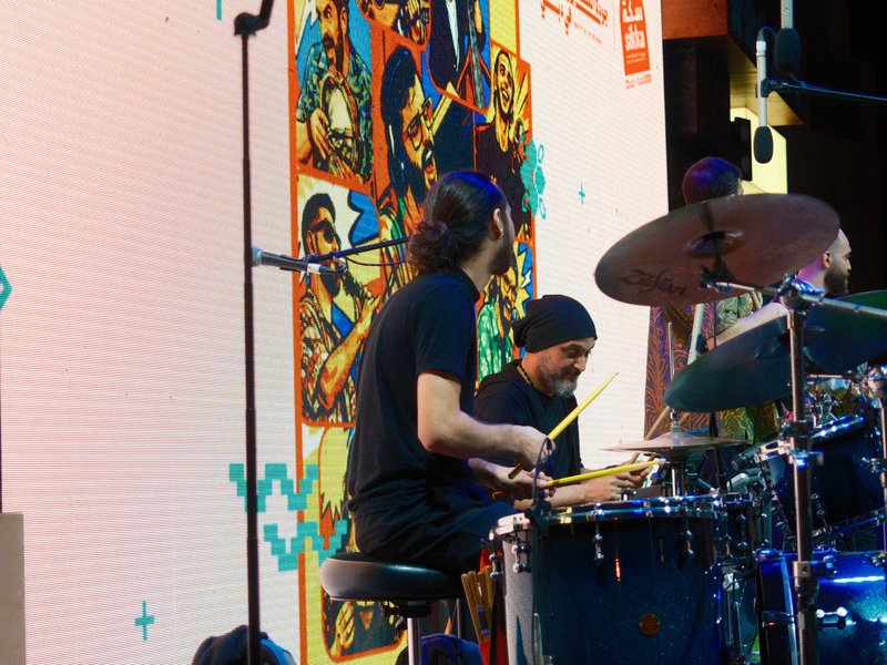 A drummer performing on stage with colorful background visuals and band members visible.