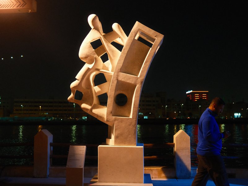 A modern sculpture illuminated at night near a waterfront, with a person walking by.