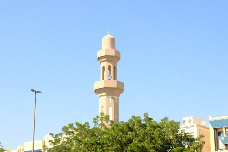 A tall minaret rises above green trees against a clear blue sky.