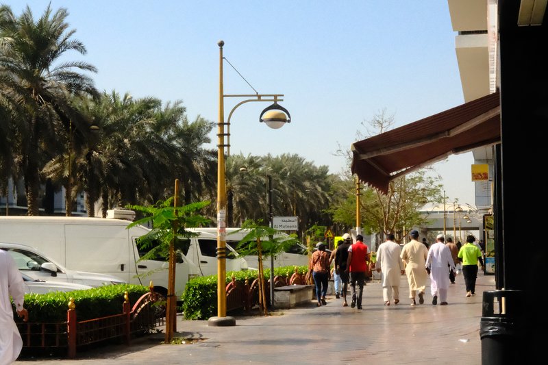A busy urban street scene with people walking along a palm-lined sidewalk under a clear blue sky.