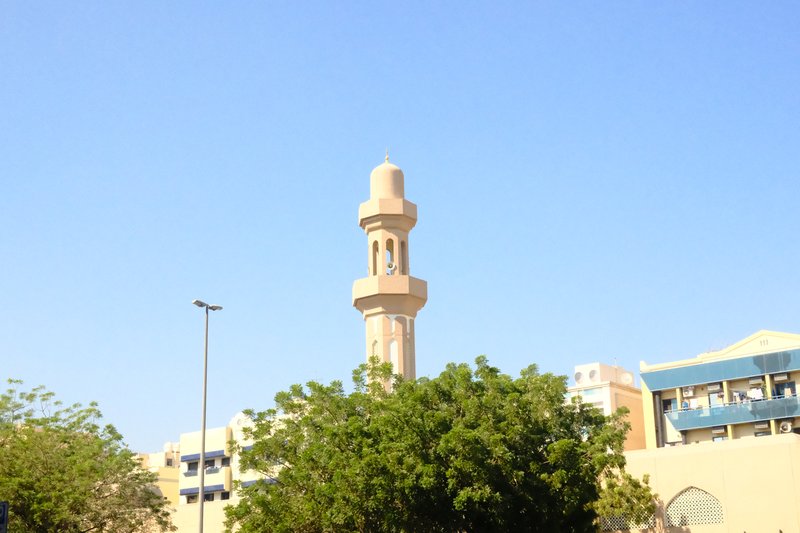 A tall minaret against a clear blue sky, surrounded by trees and buildings.