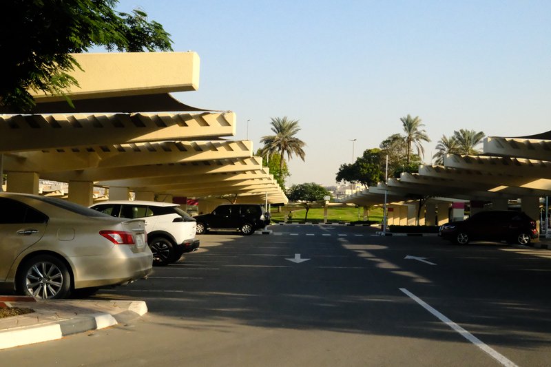 A parking lot with several cars under shaded structures and palm trees in the background.