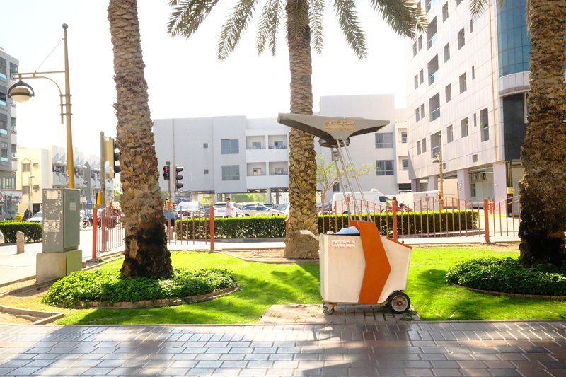 A street scene featuring palm trees and a modern trash bin in an urban setting.