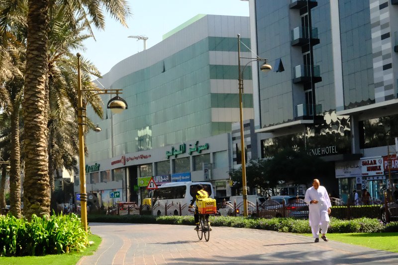 A sunny urban street scene with palm trees, buildings, and a cyclist carrying goods.