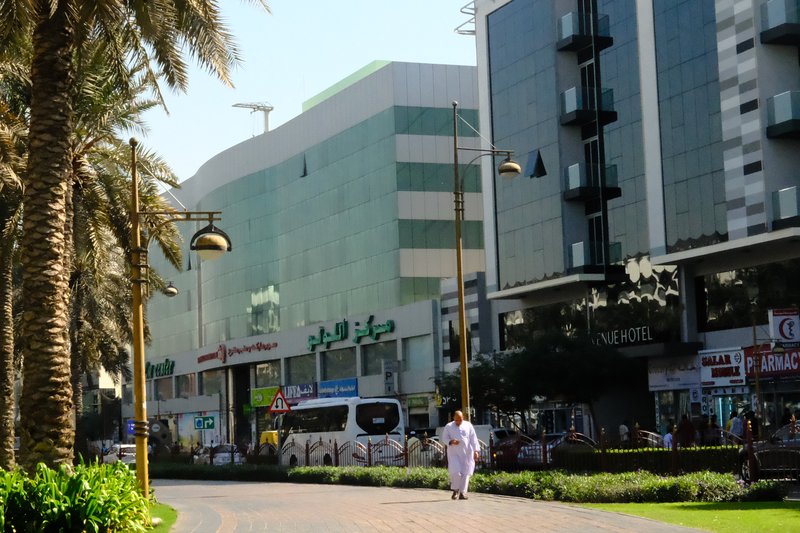 A busy urban street scene with palm trees, modern buildings, and pedestrians in a sunny atmosphere.