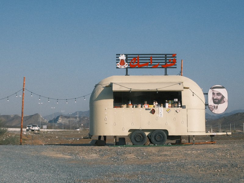 A vintage food trailer with a sign, set against a clear blue sky and distant mountains.