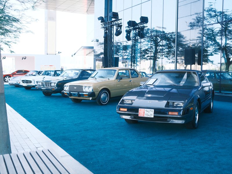 A lineup of classic cars displayed outdoors in front of a glass building with blue carpet.