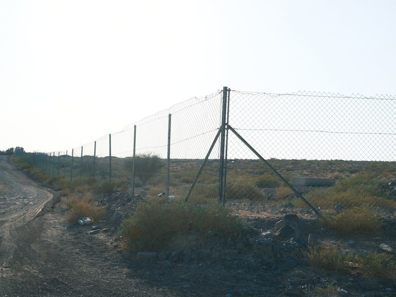 A chain-link fence along a dirt road with sparse vegetation and a bright sky in the background.