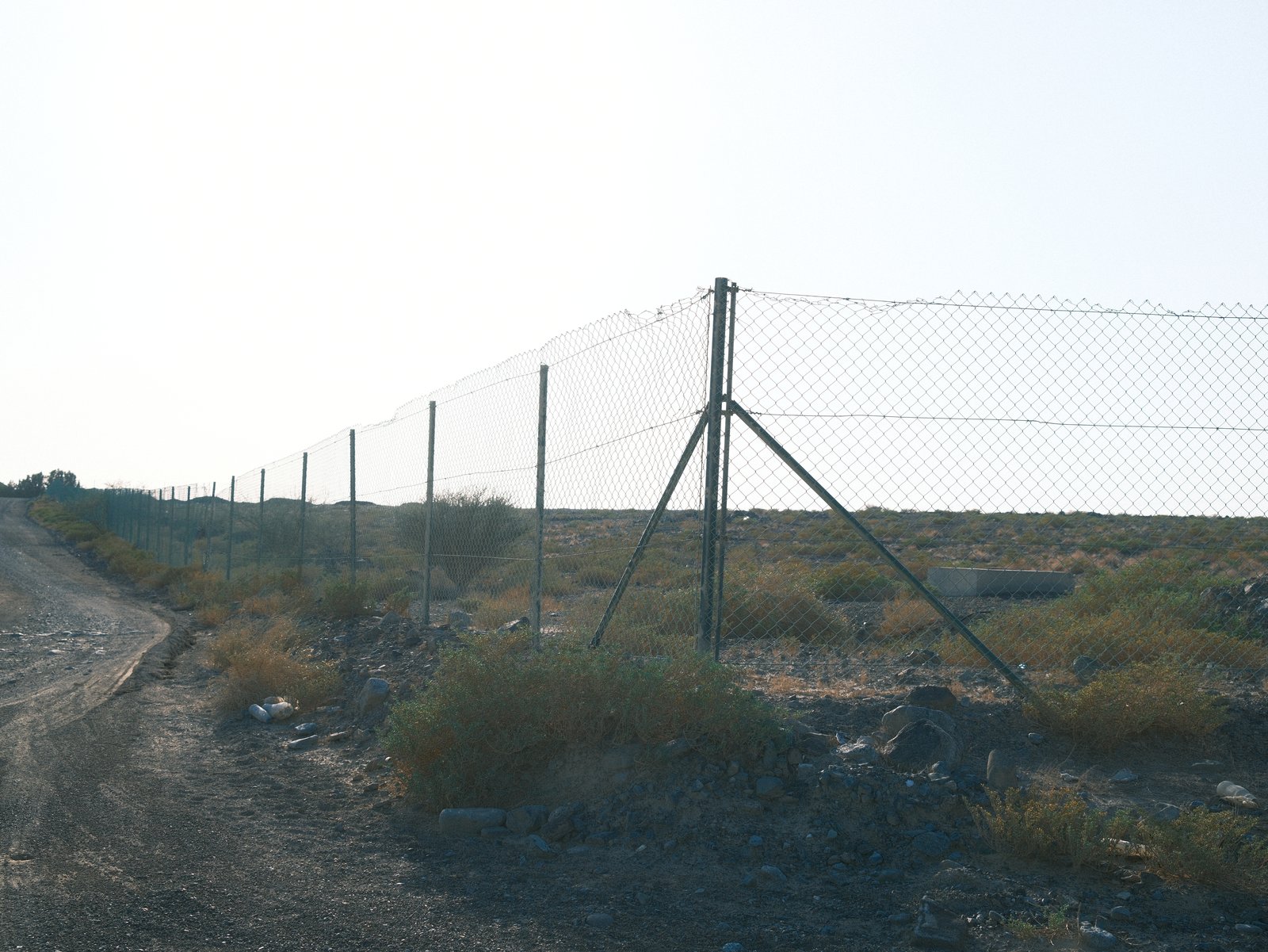 A chain-link fence along a dirt road with sparse vegetation and a bright sky in the background.