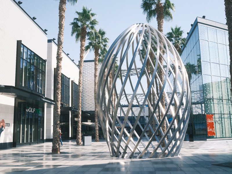 A modern metallic sculpture resembling an egg, surrounded by palm trees and contemporary buildings.