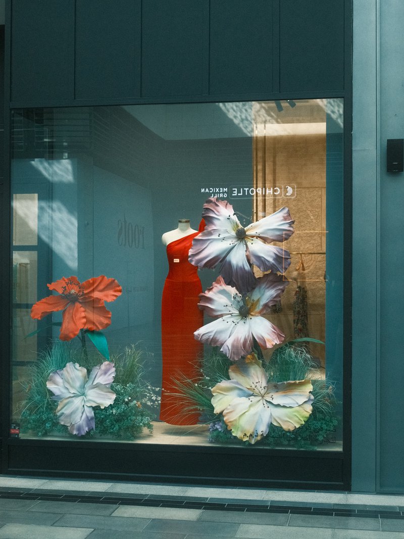 A vibrant window display featuring large flowers and a red dress on a mannequin.