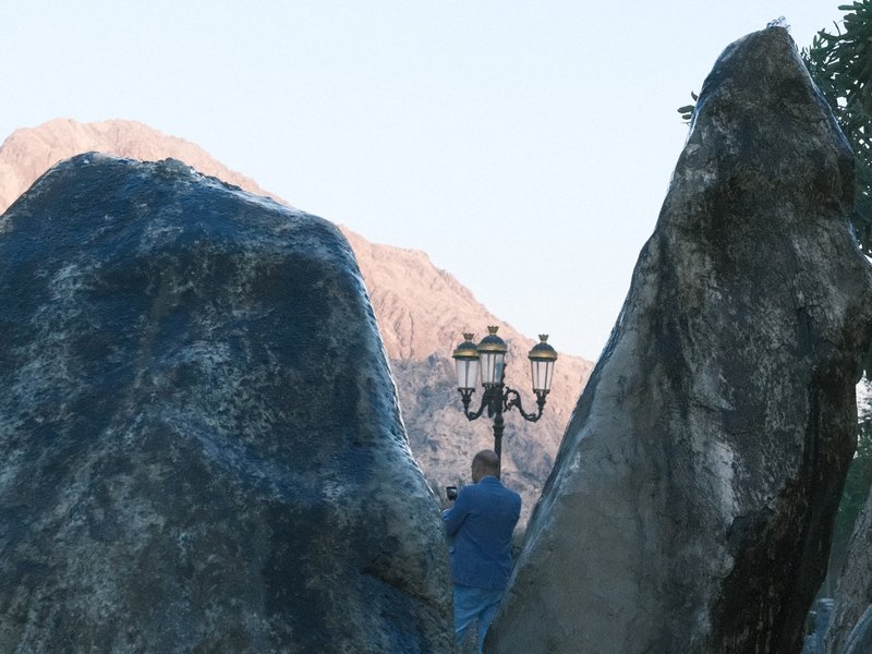 A person standing between two large rocks with a lamp post in the background and mountains in the distance.