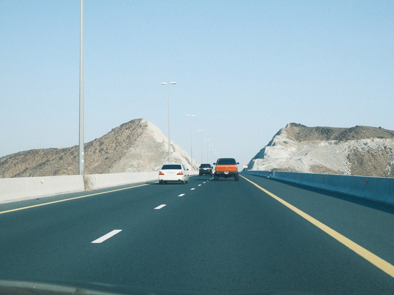 A highway scene with two cars driving towards rocky hills under a clear blue sky.