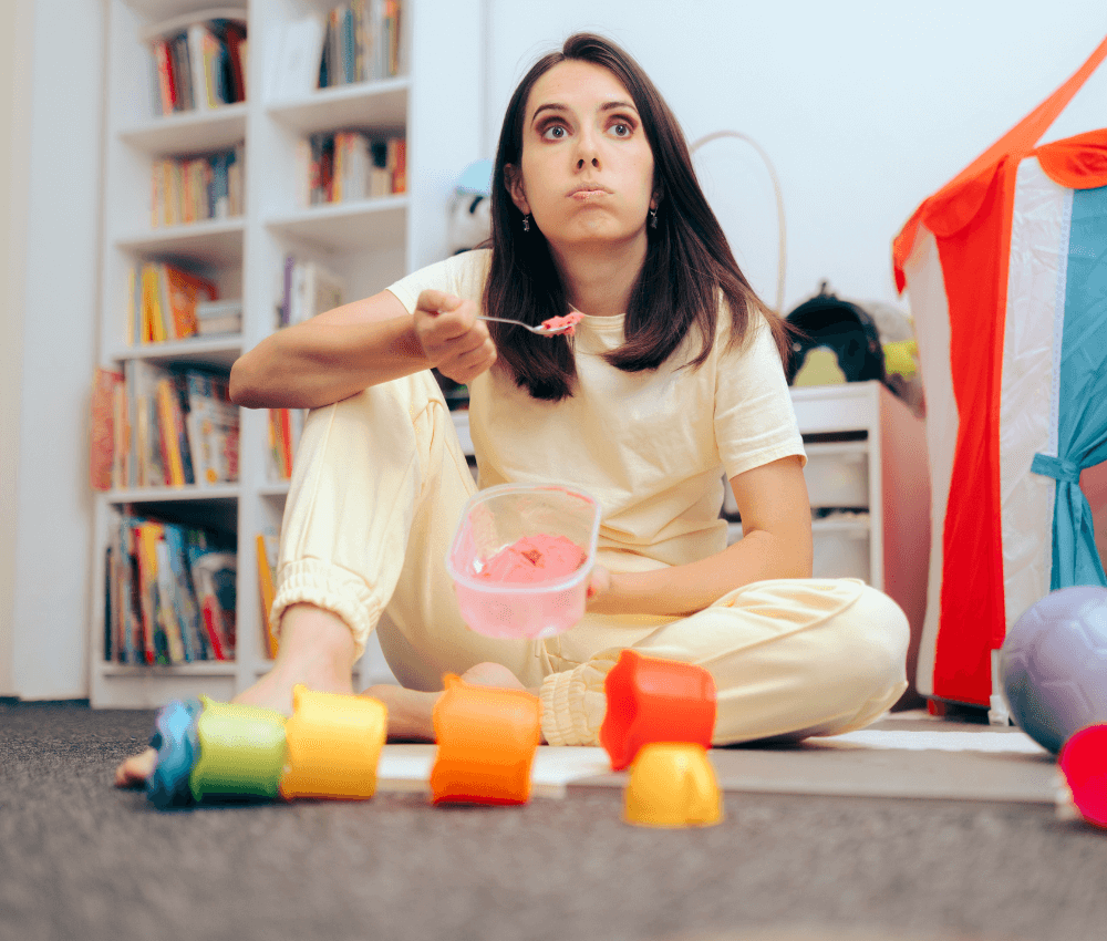 Overwhelmed nanny eating ice cream in a messy playroom, surrounded by toys and books