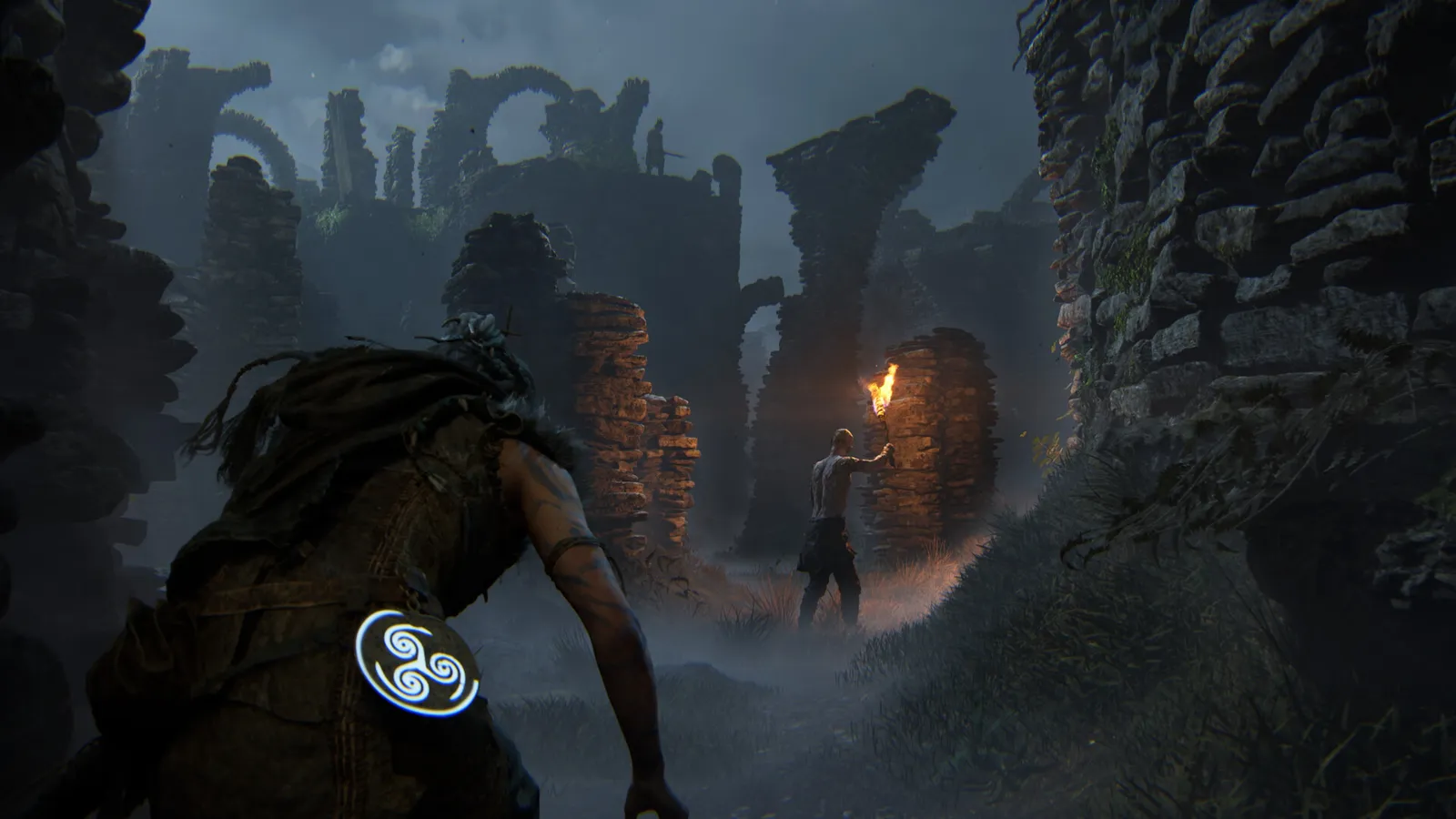 Senua sneaks between stone ruins while two shadowy creatures prowl nearby, one holding a flaming torch