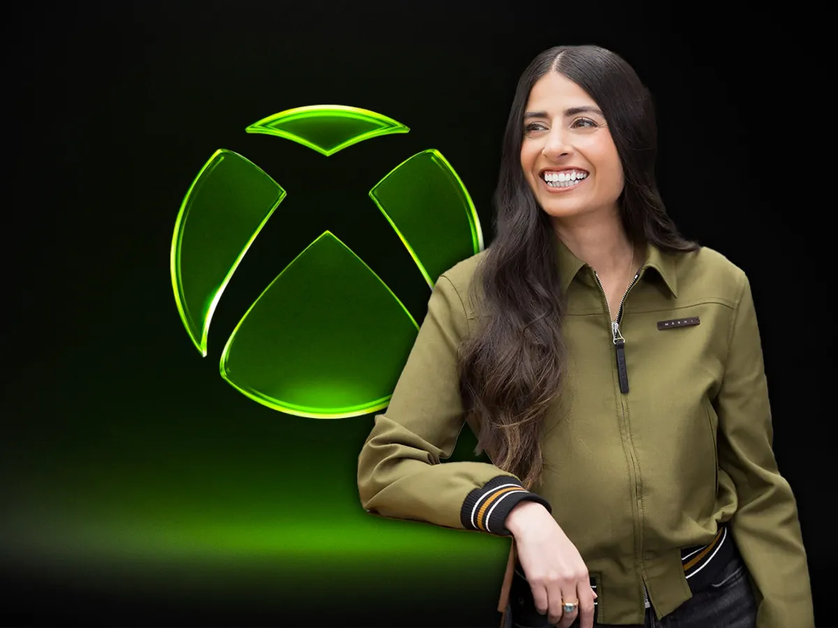 Xbox CEO Asha Sharma poses in front of the Xbox logo during a press event, smiling in a thin green jacket