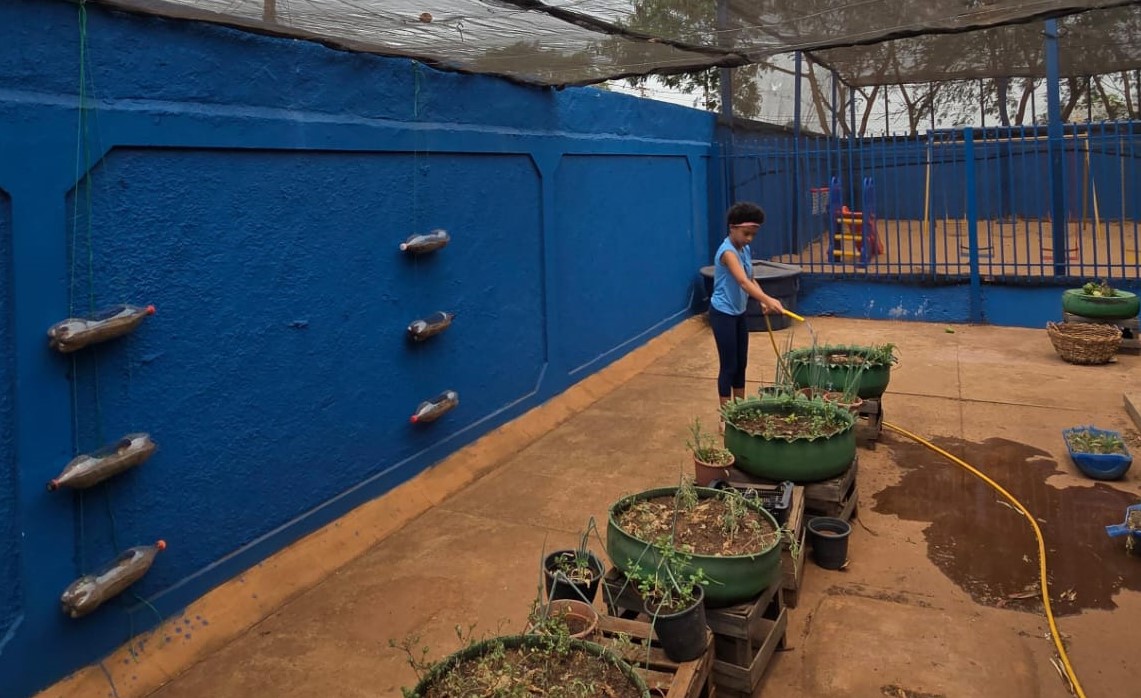 Estudante regando horta escolar com canteiros e vasos cultivados para educação ambiental