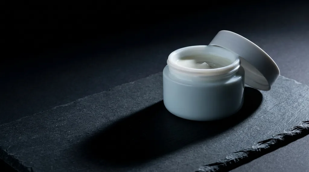 A forensic, clinical close-up of a pale white face cream jar on a dark surface, one side lit by cold laboratory light, casting a hard shadow beautiful and unsettling simultaneously, A premium white ceramic face cream jar resting on a matte dark slate surface, photographed from a 45-degree angle under cold, directional clinical lighting from one side The jars lid is half-open, cream visible inside, perfectly smooth and luxurious The harsh single-source light casts a dramatic hard-edged shadow across the surface The background transitions from pure black to a deep charcoal gray No warm tones anywhere this is clinical, slightly ominous, forensic beauty photography No flowers, no marble, no gold The aesthetic is science lab meets luxury product placement, with an undertone of unease , hyper-realistic editorial photography, 8k, sharp focus, professional color grading, NO text, NO illustration, NO cartoon, NO 3d render