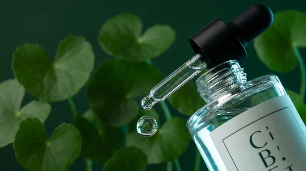 A single glass dropper bottle of clear serum with a droplet mid-fall, suspended against a dark botanical background, A single minimalist glass skincare dropper bottle with one perfectly formed droplet of clear liquid suspended mid-fall, photographed against a deep forest green botanical background with blurred centella asiatica leaves, Sony A7R IV 100mm f28 macro lens, dramatic rim lighting from the right with a cool 5600K clinical tone, visible surface tension and refraction in the droplet, luxury editorial stillness, deep green and clear glass color palette, hyper-realistic editorial photography, 8k, sharp focus, professional color grading, NO text, NO illustration, NO cartoon, NO 3d render