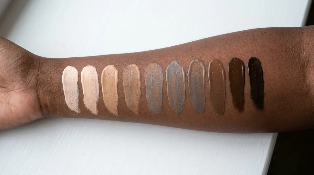 A clean white surface with a row of foundation swatches in graduating depths from porcelain to deep ebony, with the deeper swatches visibly ashy and mismatched against the skin beneath, A close-up macro shot of foundation swatches applied in a row on a human forearm with a deep brown complexion The swatches graduate from very light to deep, but several mid-to-deep shades show a visible grayish-ashy cast against the rich brown skin underneath Shot on Canon R5, 100mm f28 macro lens, from directly above Lighting soft diffused natural window light from the left, no harsh shadows, revealing true pigment accuracy Texture focus the contrast between the skins warm undertone and the cool-cast swatch pigments Mood clinical, honest, slightly melancholy  a visual document of a real problem Color palette warm mahogany skin tones contrasted with cool-gray foundation mismatches on a white background hyper-realistic editorial photography, 8k, sharp focus, professional color grading, NO text, NO illustration, NO cartoon, NO 3d render