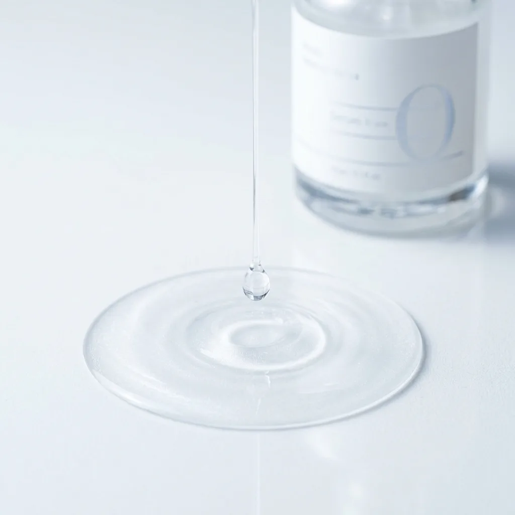 A close-up macro shot of a single drop of clear serum falling into still water, creating a perfect circular ripple, on a clean white surface with clinical cool lighting, Extreme macro close-up of a single transparent serum droplet mid-fall above a perfectly still pool of clear liquid on a pure white laboratory surface, the drop caught in suspension with a fine thread of liquid connecting it to the surface, ripples forming in a perfect circle, shot on Sony A7R IV with 100mm f28 macro lens, clinical overhead lighting at 5600K cool white, the liquid has a faint pearlescent shimmer, color palette of pure white and pale silver-blue, mood of precision and scientific integrity, hyper-realistic editorial photography, 8k, sharp focus, professional color grading, NO text, NO illustration, NO cartoon, NO 3d render