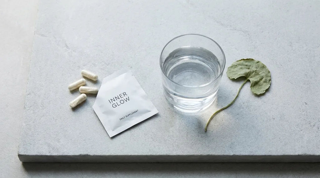 A minimalist flat lay of inner beauty supplements clear capsules and white powder sachets arranged beside a glass of water on a pale stone surface, with a single dried centella leaf as the only botanical accent, A minimalist flat lay composition of translucent collagen peptide capsules, a small sachet of white powder supplement, and a clean glass of water arranged on a cool pale grey stone surface, one dried botanical leaf as accent, shot overhead with a Sony A7R IV 50mm f28, soft diffused natural light from the left casting gentle shadows, the color palette is muted pearl white and warm stone grey with a single pale sage accent, textures sharp and tactile, serene clinical-minimal mood, hyper-realistic editorial photography, 8k, sharp focus, professional color grading, NO text, NO illustration, NO cartoon, NO 3d render