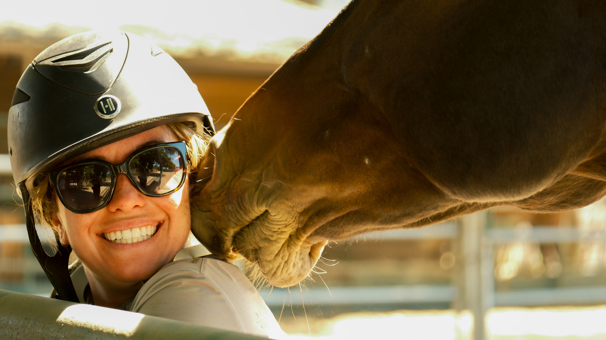 Image of a smiling woman with a helmate and sunglasses as she is getting nuzzled by a brown horse on the side of her face.