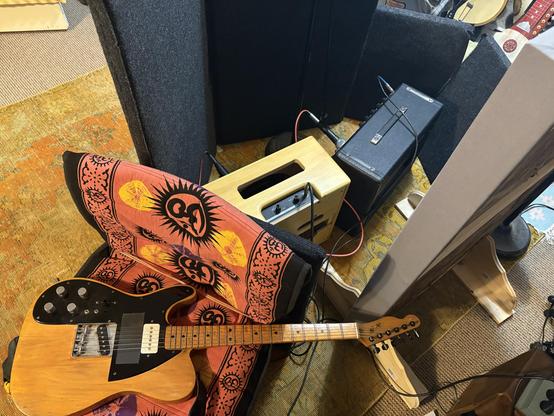 Photo of a telecaster guitar laid across a chair, which is covered by a red cloth with om symbols. Behind the guitar are two small amplifiers surrounded by sound absorbing panels. 