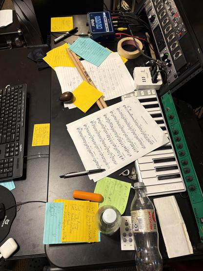 Photo of busy music studio desk with various sticky notes, sheet music, drum sticks, masking tape roll, pen, egg shaker, and other studio gear. 