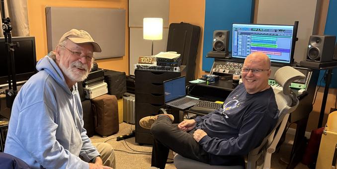 Two old white men —one w white beard, glasses, tan baseball cap and light blue hoodie, the other w a shaved head, glasses, and black long-sleeved tshirt—sitting in a home recording studio smiling at the camera. 