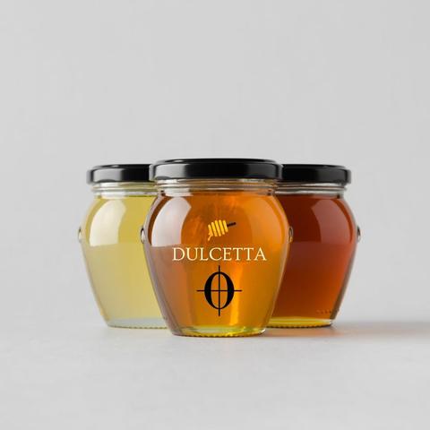 three honey jars, one in front two behind on either side. three different shades of honey color. Middle jar has printed on front a honey dipper above the word DULCETTA and a music symbol for a coda below.