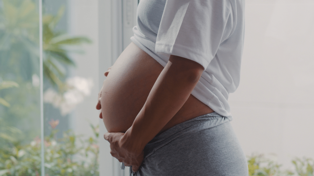 Imagem de uma mulher grávida com as duas mãos na barriga e algumas plantas de fundo