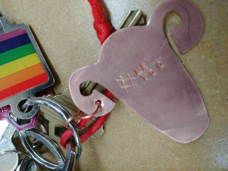 A keychain shaped like a uterus, stamped with "#METOO". A key with a rainbow inset is also seen.