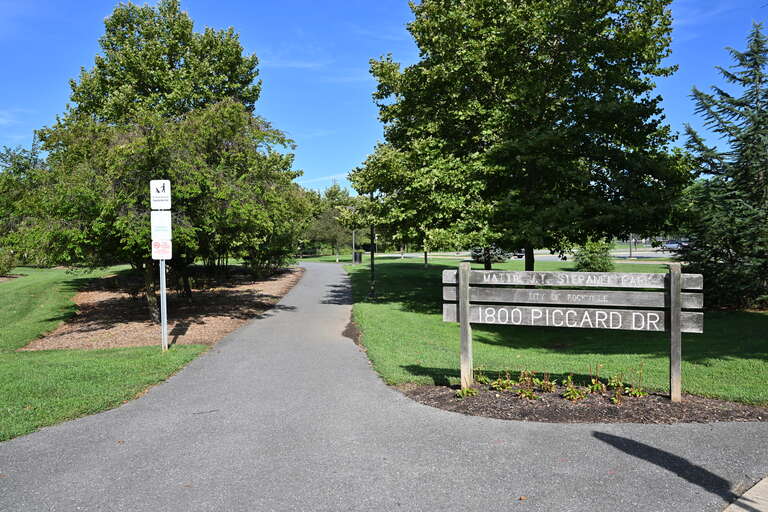 One of the entrances to Mattie J.T. Stepanek Park with a sign and a paved walkway. This park is operated by the City of Rockville.  1800 Piccard Drive, Rockville, Maryland 20850.