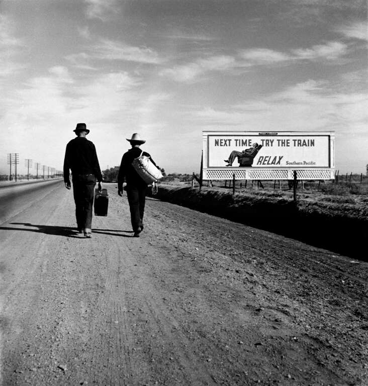 Photo shows two people walking along road near a billboard that says "Next time, try the train. Relax." Nitrate negative. 2 1/4 x 2 1/4 inches.