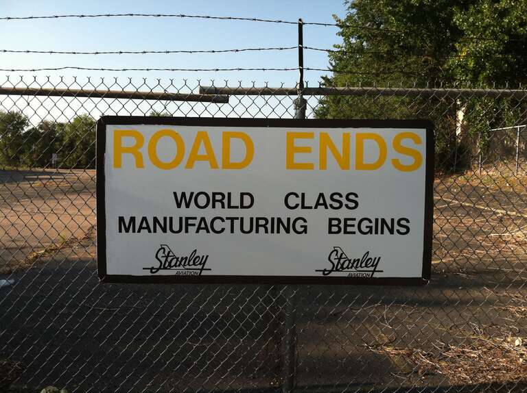 This is a sign attached to the chain link fence at the former Stanley Aviation plant at Stapleton Airport in Denver, Colorado.  The sign reads "Road Ends World Class Manufacturing Begins".  The photo was taken August 25, 2012, long after the plant