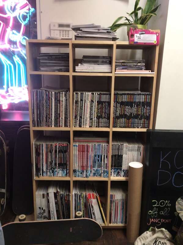 A skateboard sits in front of the bookshelves holding many awesome old issues of skate magazines from Thrasher to Strength to Slap at the Look Back Library at KCDC.