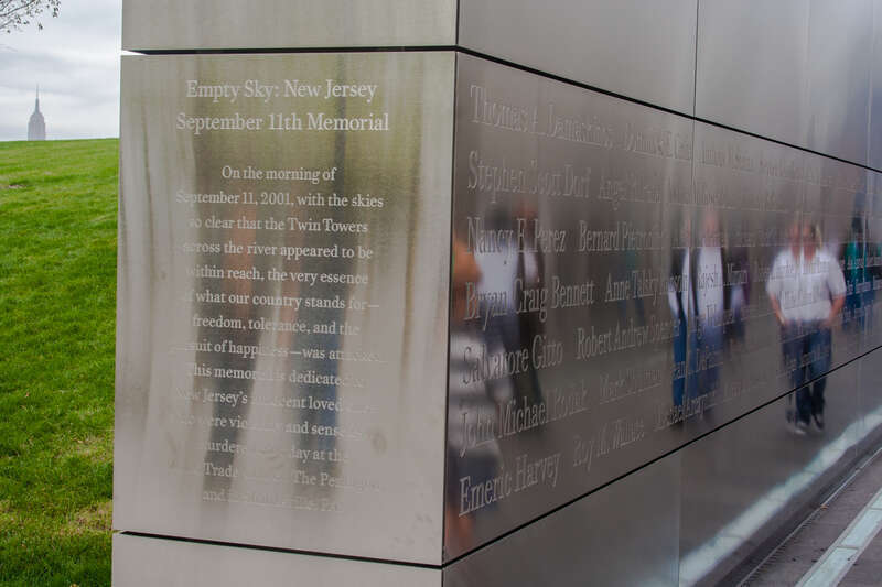 Reflections of visitors passing through the memorial.  On the left, the top of the Empire State Building is visible above the grassy hill.