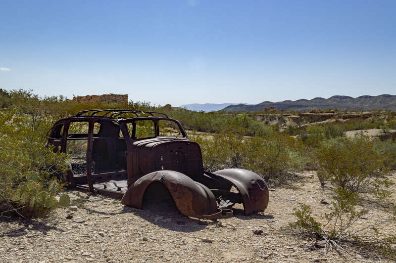 The rusted frame of a car abandoned in the Terlingua Ghost Town near the cluster of limestone buildings made by workers. The car is surrounded by scrubby cactus and creosote bushes.